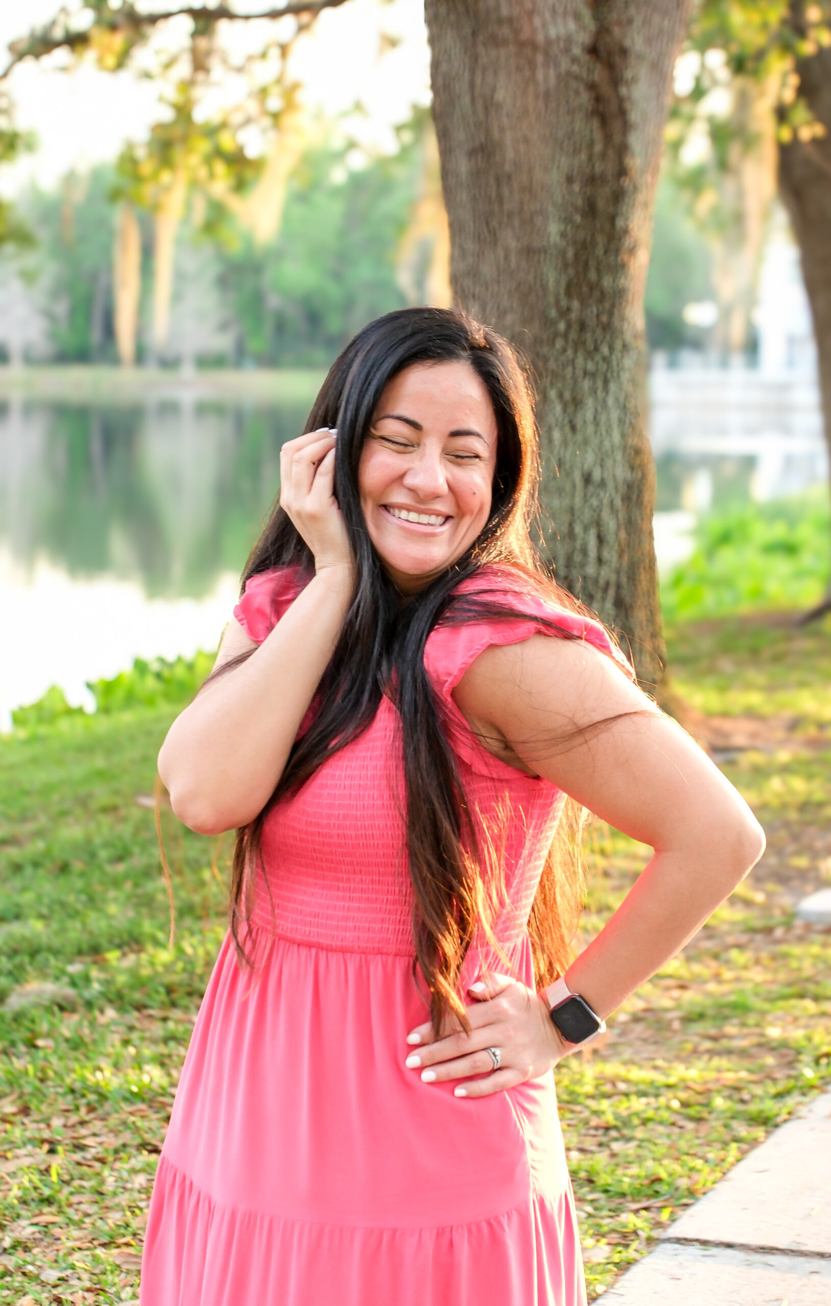A joyful woman in a coral pink dress stands outdoors near a lake, smiling with eyes closed and hand gently touching her face, radiating confidence and authenticity during a personal brand photoshoot in Celebration, Florida.